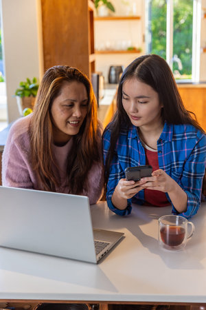 Asian mother and daughter sharing smartphone while working on laptop at kitchen table. Family, bonding, communication, modern, cozy, lifestyle, hydrationの写真素材