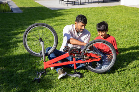Asian father and son kneeling in backyard adjusting red bicycle chain near helmet and scooter....