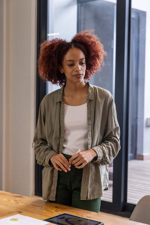 African american woman standing beside table and glass door in office using touchscreen tablet....