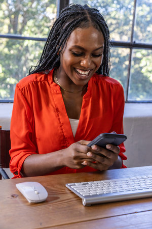 African American woman smiling while checking smartphone at desk by window with keyboard and mouse....