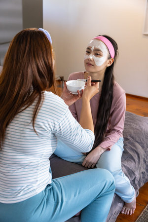 Diverse female friends applying white facial mask cream on bed using ceramic bowl and brush....