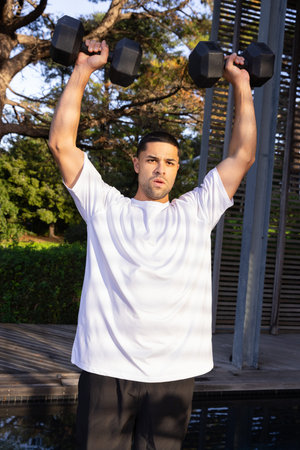 Man lifting hexagonal dumbbells overhead on wooden deck beside reflecting water under metal pergola. Fitness, outdoor gym, wellness, strength training, natural environment, active lifestyle, healthy livingの写真素材