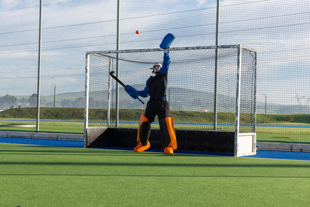 Male field hockey goalkeeper with helmet leaping and blocking orange ball on green turf at goal. Athlete, sports, action, safety, protection, competition, outdoorの写真素材
