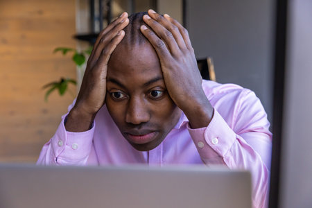 African American man holding head while staring at laptop screen at office desk with plant shelf....