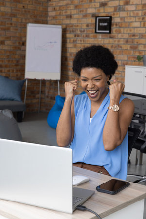 African American woman cheering on laptop at wooden desk in modern office lounge with flipchart....