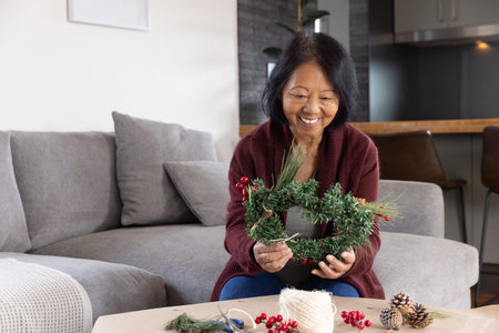 Senior Asian woman sitting in living room crafting wreath with pine cones, red berries, copy space....