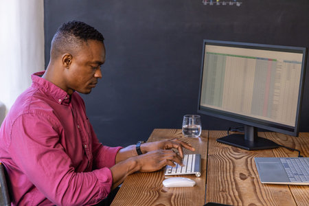 African American man typing spreadsheet data on wireless keyboard at wooden desk in home office....