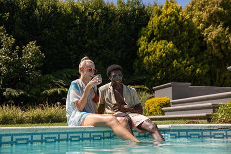 Diverse couple sitting on tiled pool edge dipping legs in water and sipping drinks wearing shades....