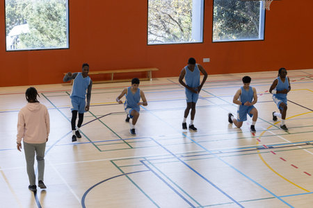 Diverse male basketball team in blue jerseys performing lunges on gym court under coach supervision....