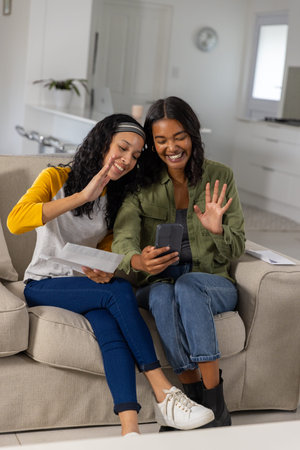 Sisters sitting on sofa waving, holding smartphone and letter by kitchen island with laptop. Family, companionship, home, modern, communication, lifestyle, cozyの写真素材