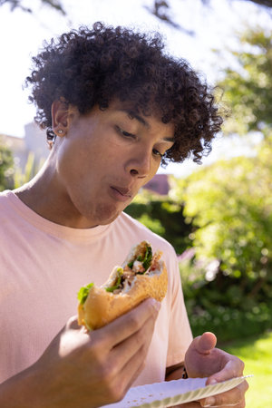 Sesame sandwich is resting on paper plate in sunlit backyard garden with lush foliage. Vibrant,...