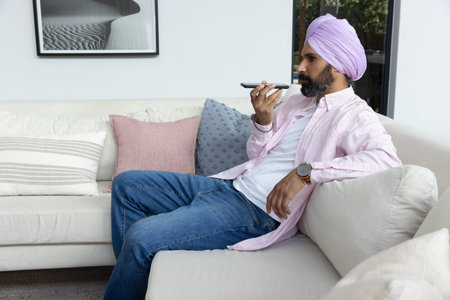 Mid adult Indian man holding smartphone voice command sitting at home with cushions, copy space....