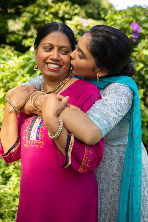 Indian mother and daughter hugging and kissing cheeks in garden with pink embroidered kurta....