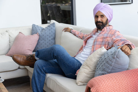 Indian man leaning back on sectional sofa at home, wearing lavender turban, wristwatch, copy space....