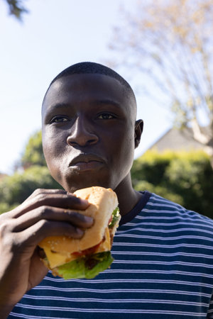 African American man standing in backyard holding and eating big sandwich with lettuce on sunny day....