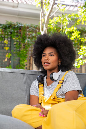 African American woman sitting on sofa in garden patio wearing yellow overalls with headphones....