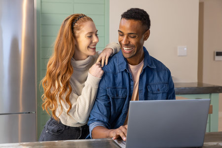 Diverse couple leaning on stone countertop in kitchen near green cabinets while using silver laptop....