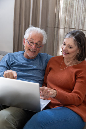 Senior couple leaning forward toward silver laptop on neutral-tone sofa in home living room....