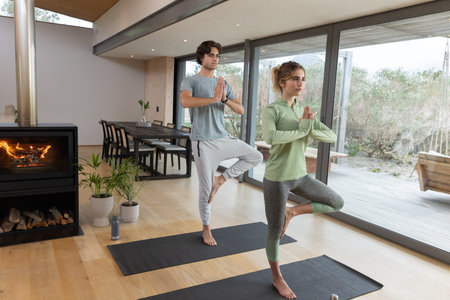 Couple practicing yoga pose in athletic wear on black mats in living area with wood stove....