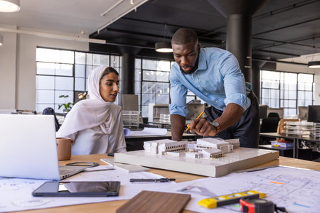 Diverse coworkers examining architectural scale model at modern office with blueprints and laptop. Collaboration, teamwork, professional, design, innovation, urban, creativeの写真素材