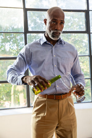 Senior African American man at window in blue shirt pouring wine from green bottle into glass....
