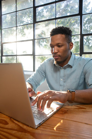 African American man working at wooden table by grid window on silver laptop, wearing leather watch....