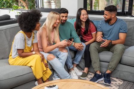 Diverse friends sitting on gray sectional sofa at home, sharing smartphone and game controller. Group, living, couch, lifestyle, modern, cozy, casualの写真素材