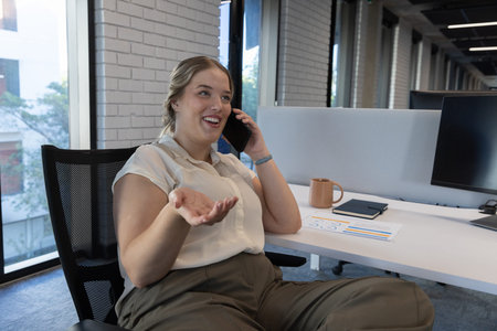 Woman sitting in mesh chair, holding smartphone and gesturing at white desk with mug in office....