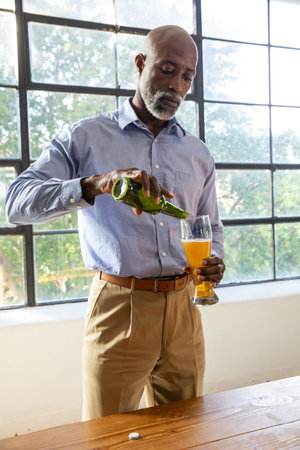 Senior African American man pouring beer from green bottle into pilsner at table wearing blue shirt....