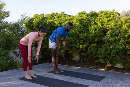 Diverse friends bending forward on wooden deck, practicing yoga on black mats in athletic clothing....