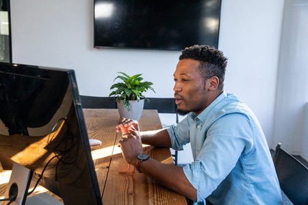 African American man leaning forward, speaking at wooden table with monitor, blue shirt, copy space....