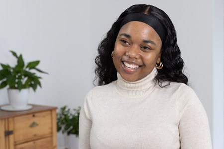 African American woman smiling and looking left in room wearing cream top black-headband gold hoops....
