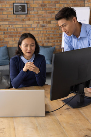 Asian team in blue shirt navy sweater collaborating at small office desk leaning to monitor laptop....