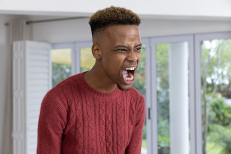 African American man wearing red cable knit sweater shouting and leaning at home near sliding doors....