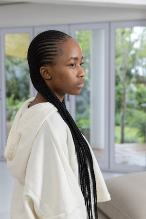 African American woman standing at home wearing hoodie showing braids beside glass doors....
