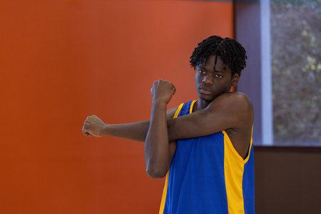 African American teen in blue-yellow jersey stretching arm across chest near window, copy space. Athlete, gym, court, training, conditioning, focus, determinationの写真素材