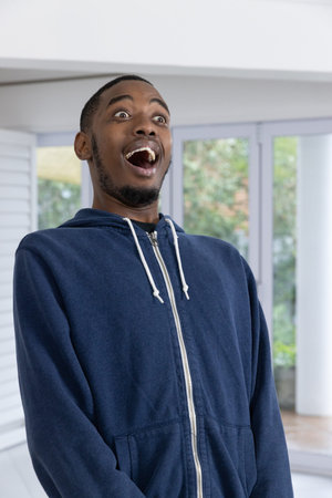 African American adult man standing in navy hoodie by glass doors and column, showing astonishment....