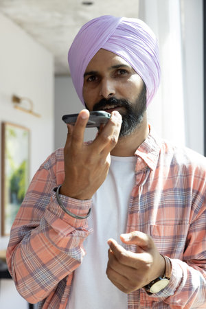 Man speaking on smartphone speaker while standing near window under brass picture light. Technology,...