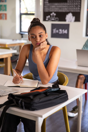 Teen girl student sitting at desk in classroom writing in notebook with pencil and laptop....