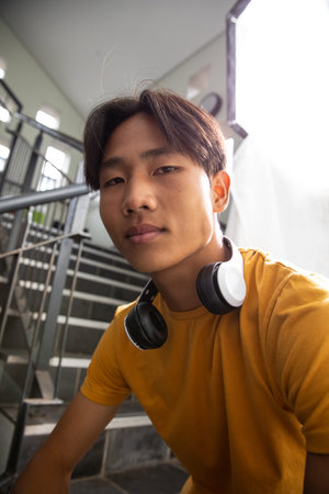 Asian teenage boy sitting on stairs in stairwell wearing yellow tee and headphones next to softbox....