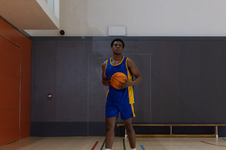 African American man holding orange basketball, standing on hardwood court in gym in blue jersey....