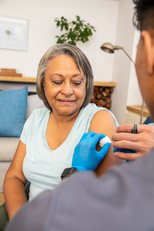 Senior woman sitting on sofa at home while provider preparing vaccine with syringe and cotton swab....