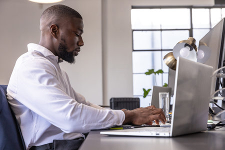 African American man typing on laptop at modern office desk with monitor and notebook, copy space....