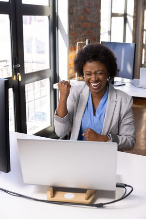 African American woman celebrating success using laptop stand and smartphone at office windows....
