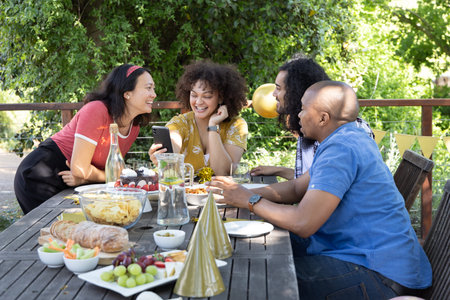 Diverse friends celebrating around picnic table on deck with snacks, smartphone, gold party hats....