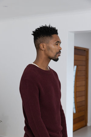 African American man standing facing right in hall wearing maroon sweater showing wooden door....