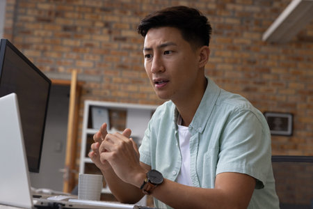 Asian man sitting at desk gesturing in meeting on laptop and monitor with mug, copy space....