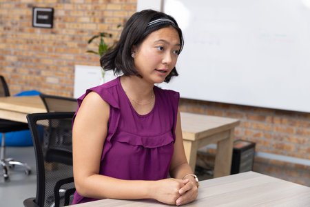 Asian woman leaning at table in meeting room wearing purple blouse with whiteboard, potted plant....