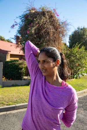 Asian Indian woman stretching in front of red-tiled house wearing purple top over pink sports bra....