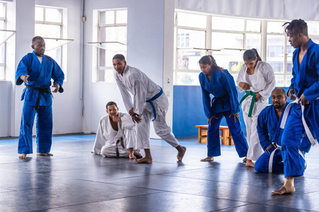 Diverse martial arts class practicing grappling drills on padded mats in dojo with gis and belts....